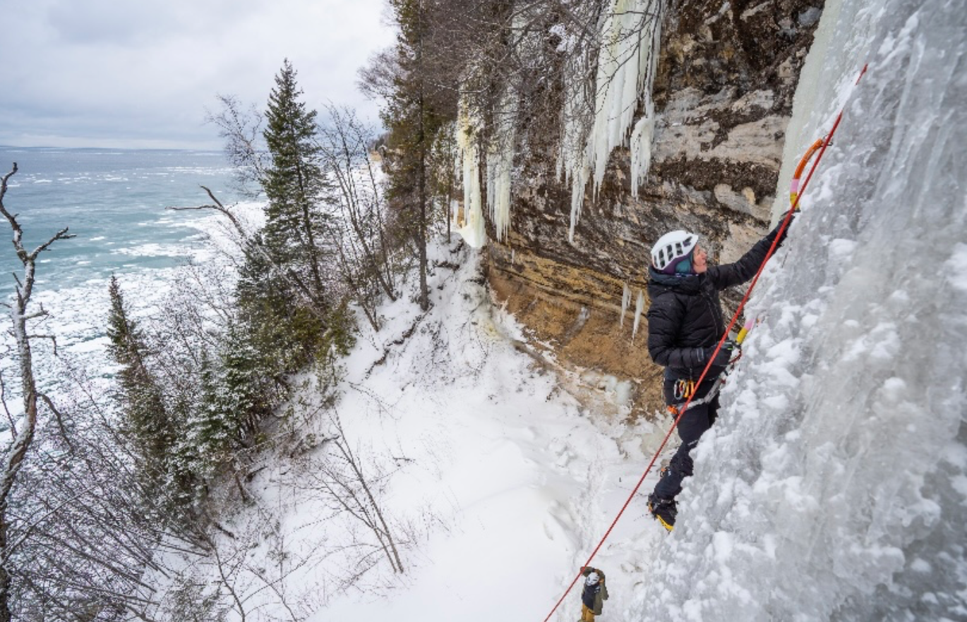 A person climbs a frozen waterfall using picks and helmets. The landscape is snowy and frozen.